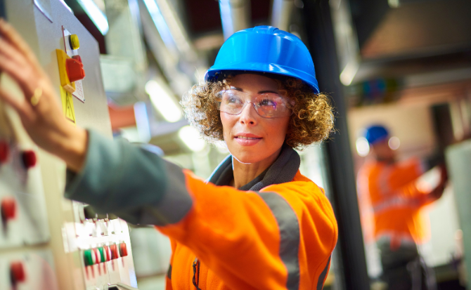 Worker in an orange safety jacket and blue hard hat operating a control panel in an industrial setting, with another worker in the background