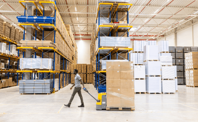 A warehouse worker, a man wearing a grey shirt and dark trousers, uses a yellow electric pallet jack to move a stack of brown cardboard boxes down an aisle