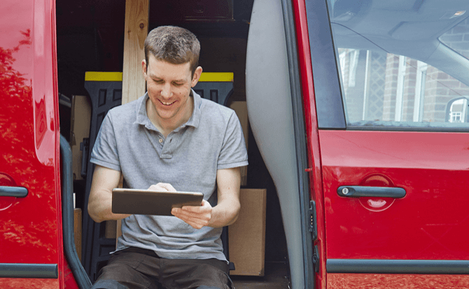A smiling male service technician, wearing a grey shirt, sits on the threshold of a red delivery van, intently using a tablet