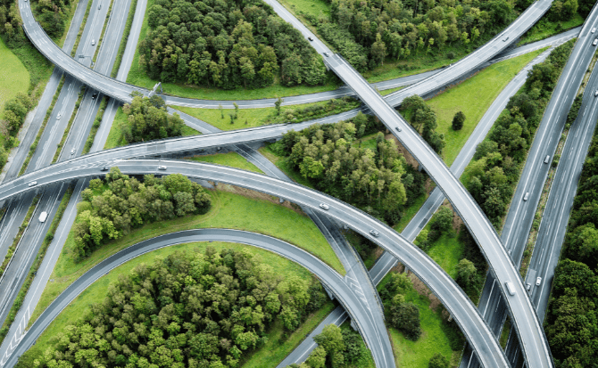 An aerial, high-angle view captures a vast and complex highway interchange, featuring multiple elevated ramps curving through a landscape of lush green trees.