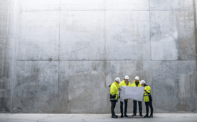 Construction site scene with four people in safety gear looking at plans, standing in front of a vast concrete wall