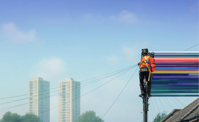 A utility worker wearing a safety harness, hard hat, and high-visibility vest is positioned on a utility pole, working on overhead power lines.