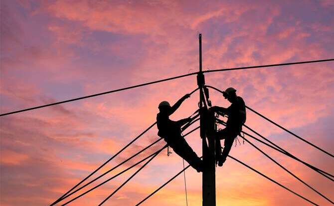 A striking low-angle photo of two technicians, silhouetted against a vibrant orange, pink, and purple sunset, working on the lines and equipment atop a tall utility pole.