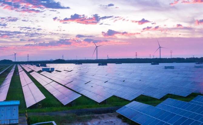 A renewable energy landscape featuring numerous solar arrays in the foreground and multiple wind turbines in the background, set against a vibrant sunset.
