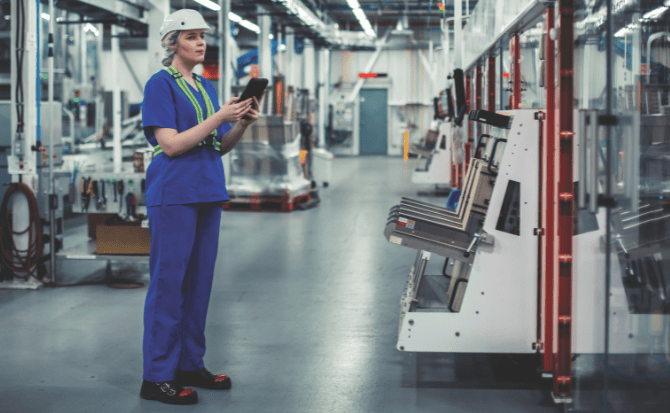 Female factory worker in a blue uniform and white hard hat holding a tablet and inspecting machinery on the factory floor.