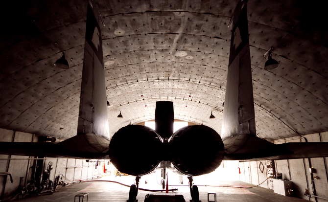 Twin-engine fighter aircraft viewed from the back in an arched-roof hangar.