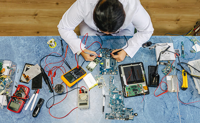 A person in a white lab coat repairing a circuit board at a workbench.