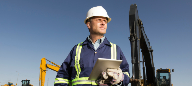 Construction asset management professional using a tablet on-site to monitor heavy equipment and ensure efficient resource utilization.