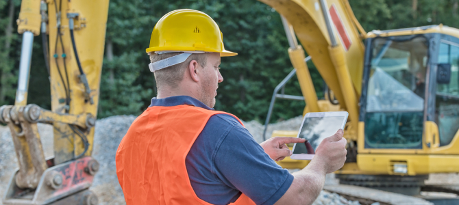 Construction worker using tablet on-site with excavator in background, leveraging Construction ERP software for real-time project management.