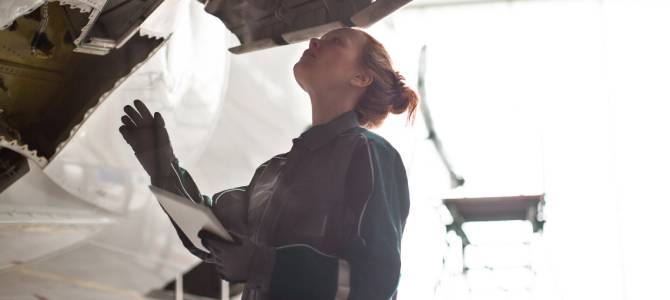 A technician in protective gear inspects the underside of an aircraft while using defense asset management software on a tablet to document and manage maintenance tasks.