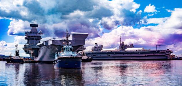Two large naval ships docked at a harbor with smaller tugboats assisting, under a partly cloudy sky reflecting blue and purple hues on the water.