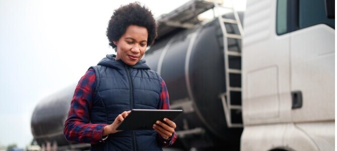  Person using supply chain software on a tablet standing in front of a large truck with a tanker trailer